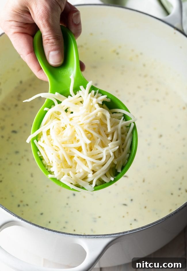 Ultimate Cheesy Chicken Alfredo Bake 5 Hand holding a green measuring cup filled with shredded mozzarella cheese, ready to be added to the sauce.