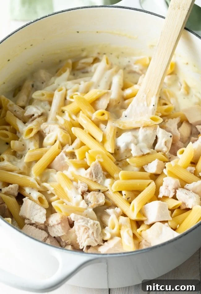 Ultimate Cheesy Chicken Alfredo Bake 6 Large cooking pot showing penne pasta, cooked chicken chunks, and a creamy Alfredo sauce being stirred with a wooden spoon.