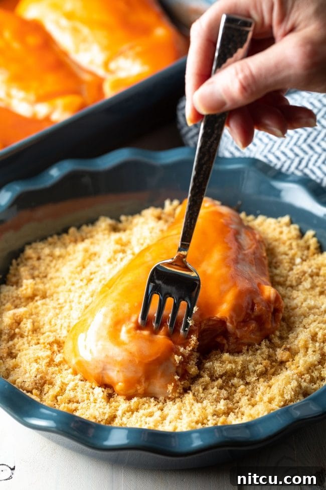 A chicken breast being generously coated with panko breadcrumbs in a pie dish.