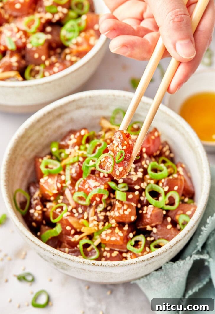 A hand gracefully holds a piece of perfectly cubed raw tuna with chopsticks, with more delectable Ahi Poke blurred in the background, showcasing its freshness and vibrant color.