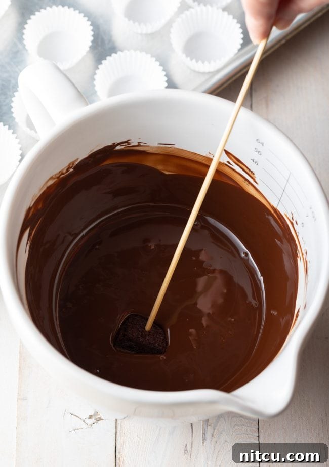 A mini chocolate cake being dipped into a bowl of melted chocolate with a wooden skewer.