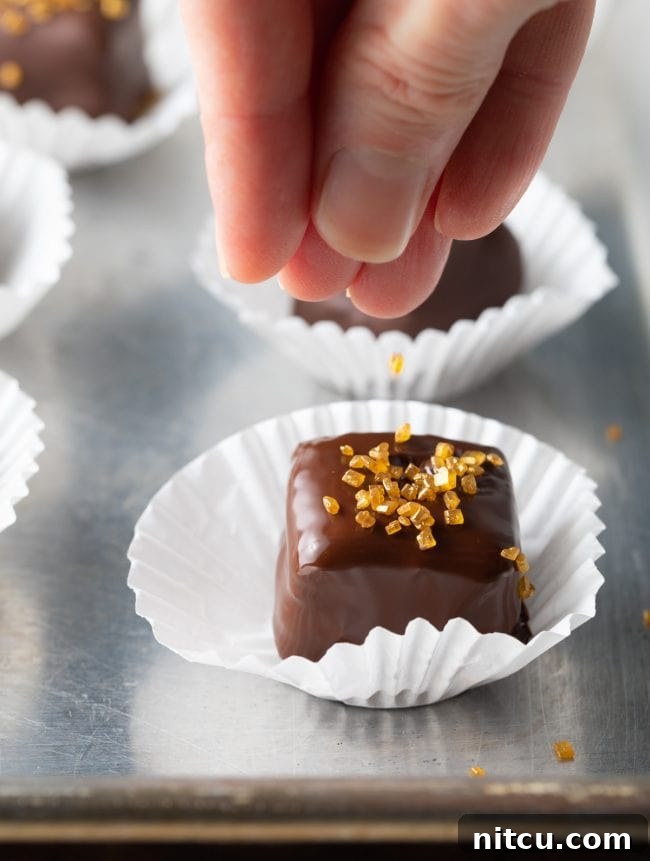 A chocolate petit four being placed onto a paper liner, adorned with colorful sprinkles before the chocolate sets.