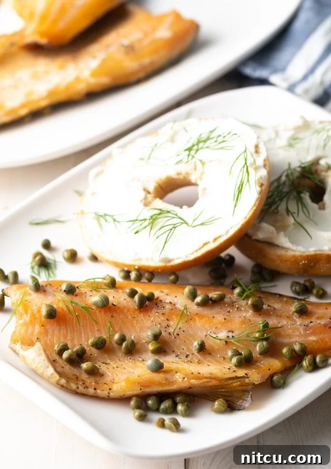 A perfectly smoked trout fillet on a white plate, garnished with capers, alongside a freshly sliced bagel.