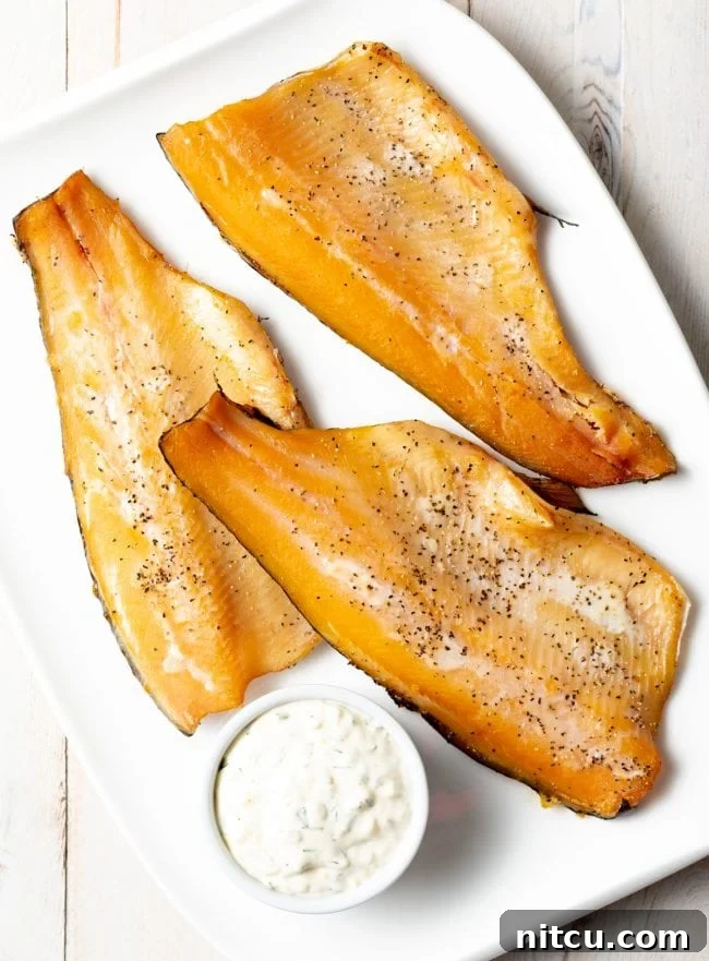 Three golden-brown smoked trout fillets arranged artfully on a white plate next to a small bowl of dipping sauce.
