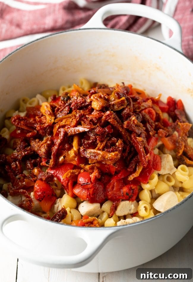 Luscious Tuscan Chicken Pasta 6 White cooking pot with chunks of cooked chicken and raw macaroni pasta noodles, topped with roasted red peppers and sun dried tomatoes.