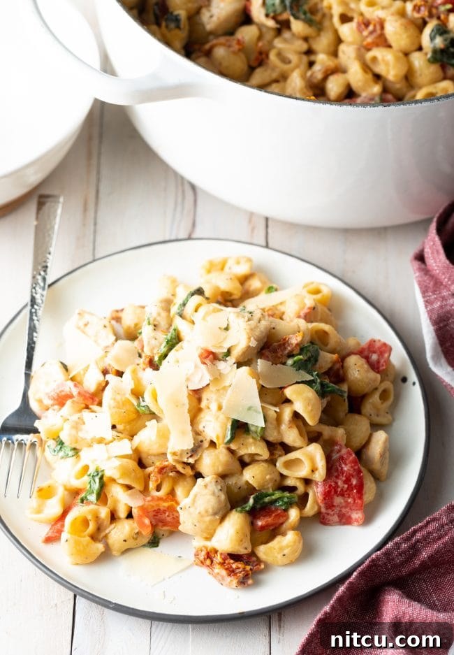 Luscious Tuscan Chicken Pasta 9 White plate with a serving of creamy chicken spinach pasta with red peppers, sun dried tomatoes, and topped with shaved parmesan cheese.
