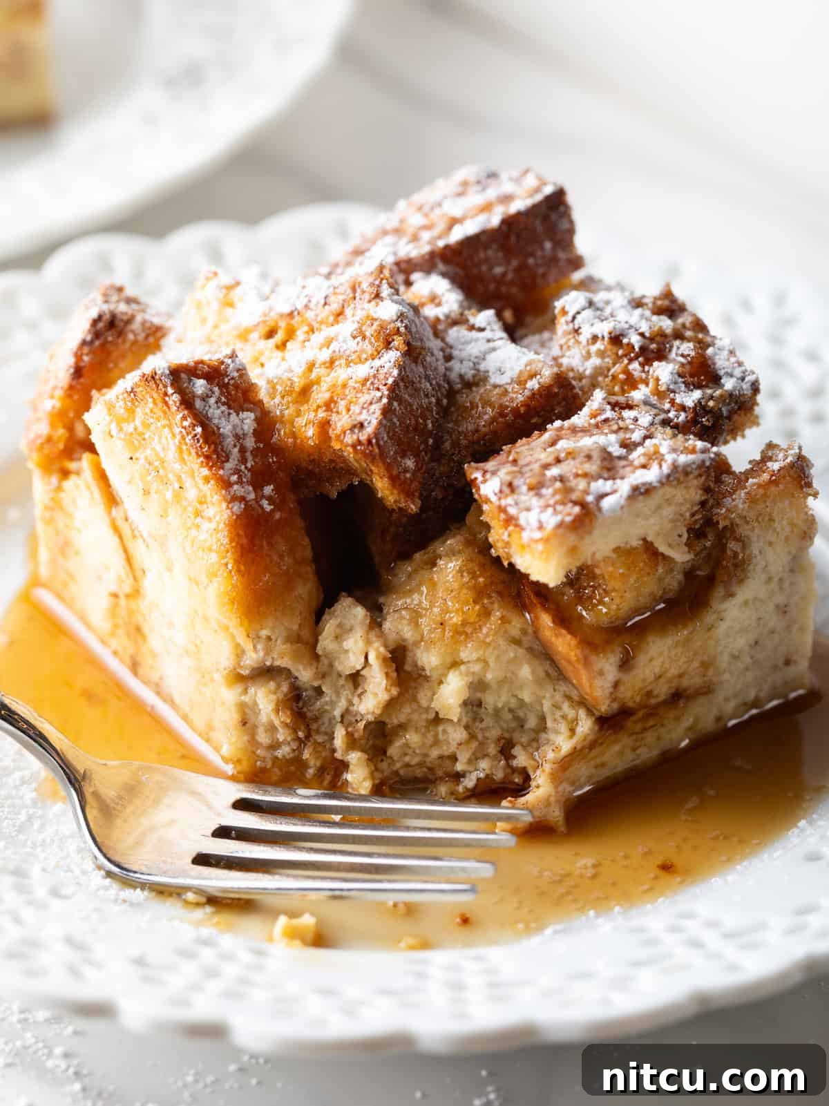 A serving of French toast casserole overnight recipe on a white plate drizzled with syrup, with a fork by its side.
