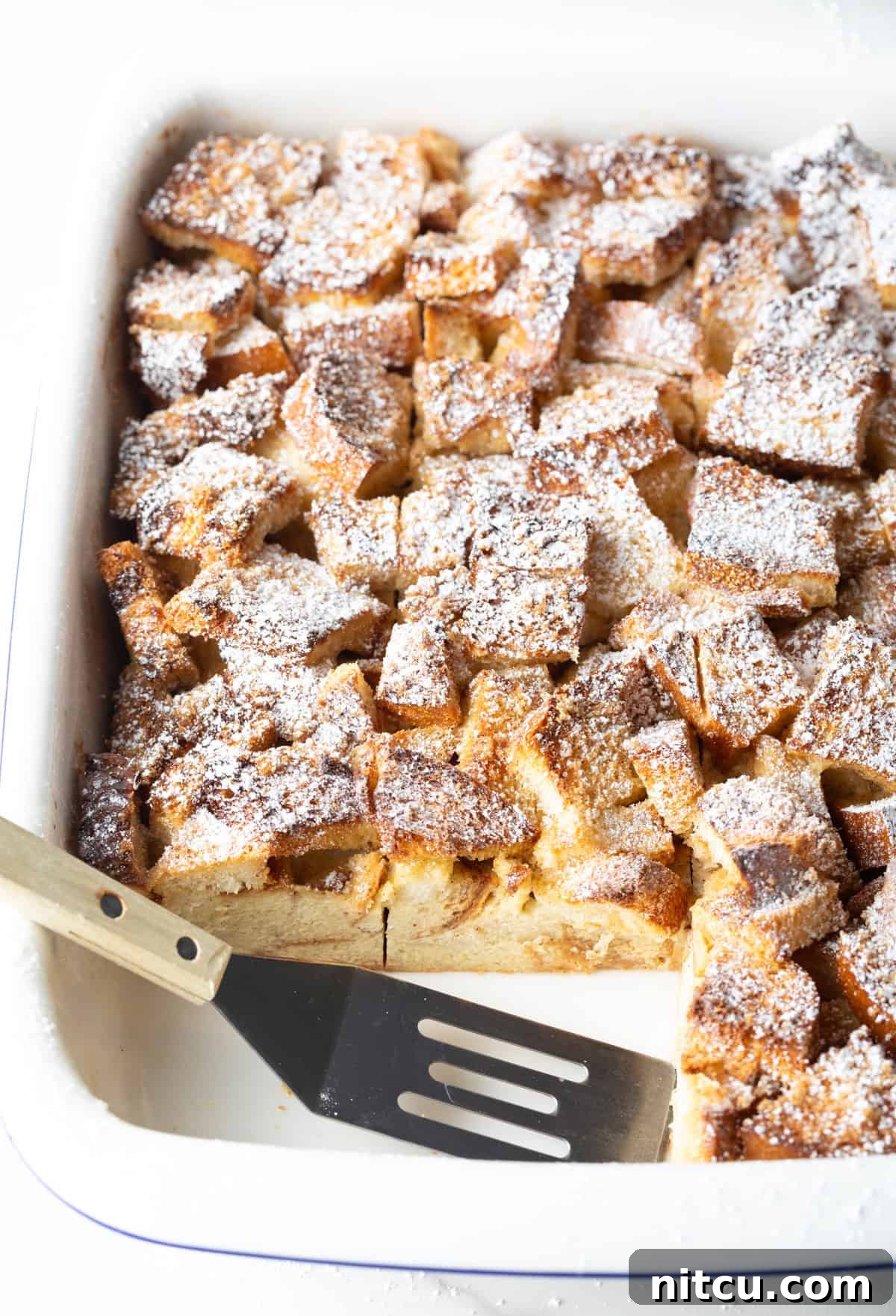 A white baking dish filled with overnight French toast breakfast casserole, with a metal spatula having taken two slices out.