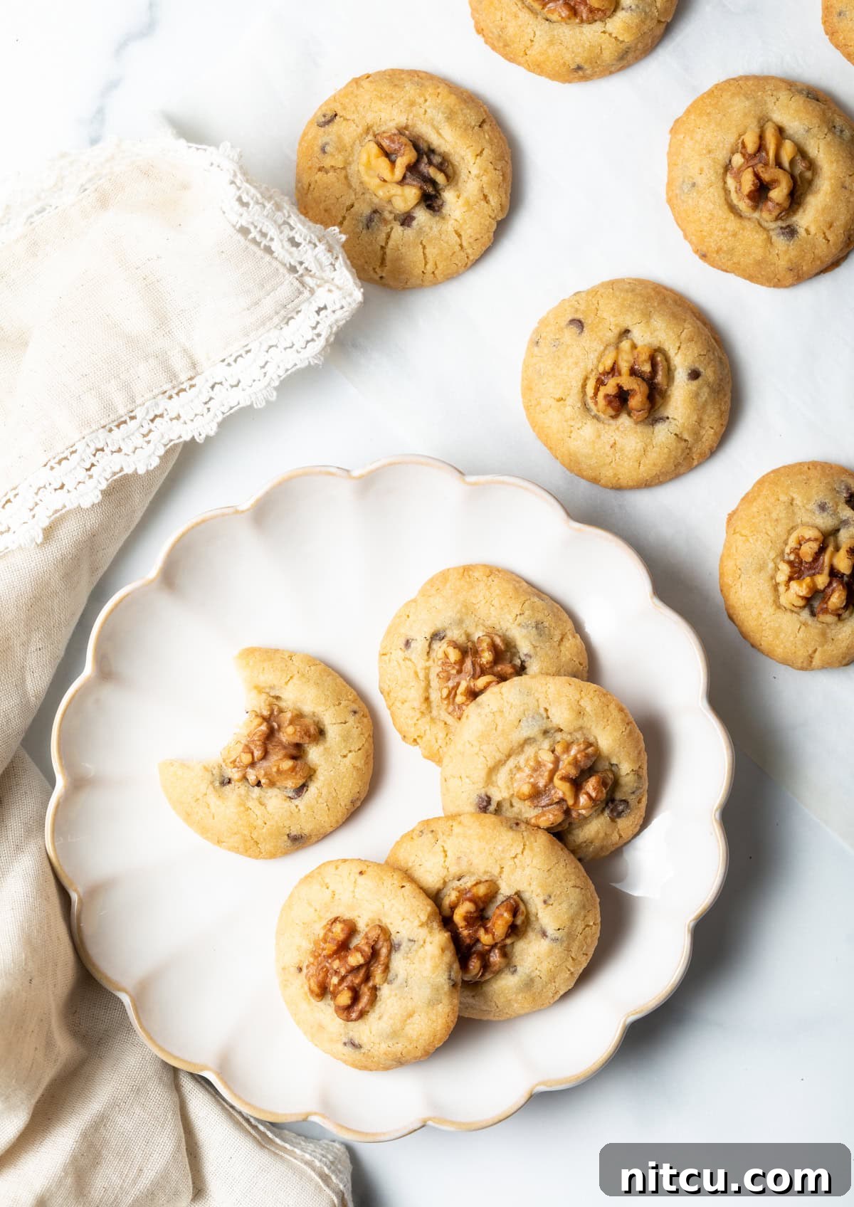 Overhead shot of chocolate chip and walnut cookies stacked in a semi-circle on a white plate, with one cookie that has a bite taken out of it.