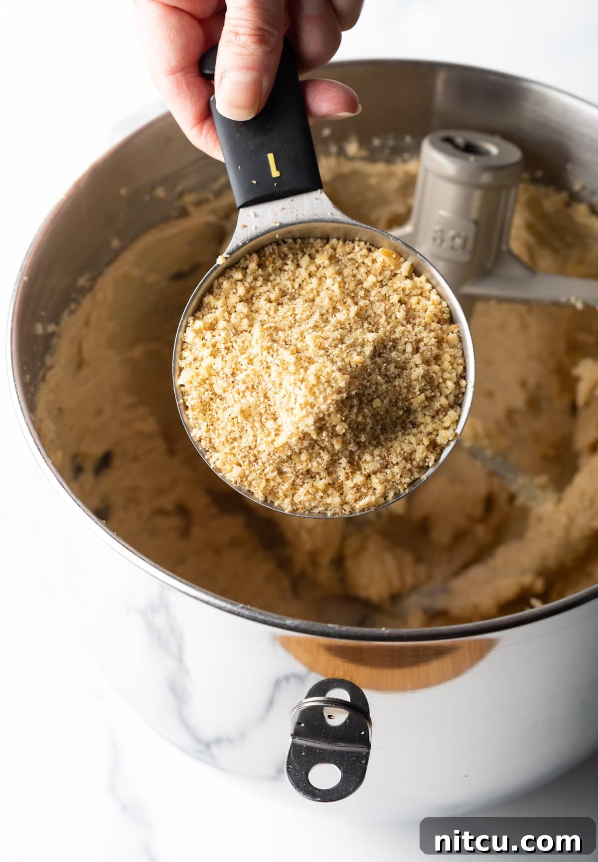 A cup of ground walnuts is being held over a stand mixer, ready to be added to the chocolate walnut cookie mix.
