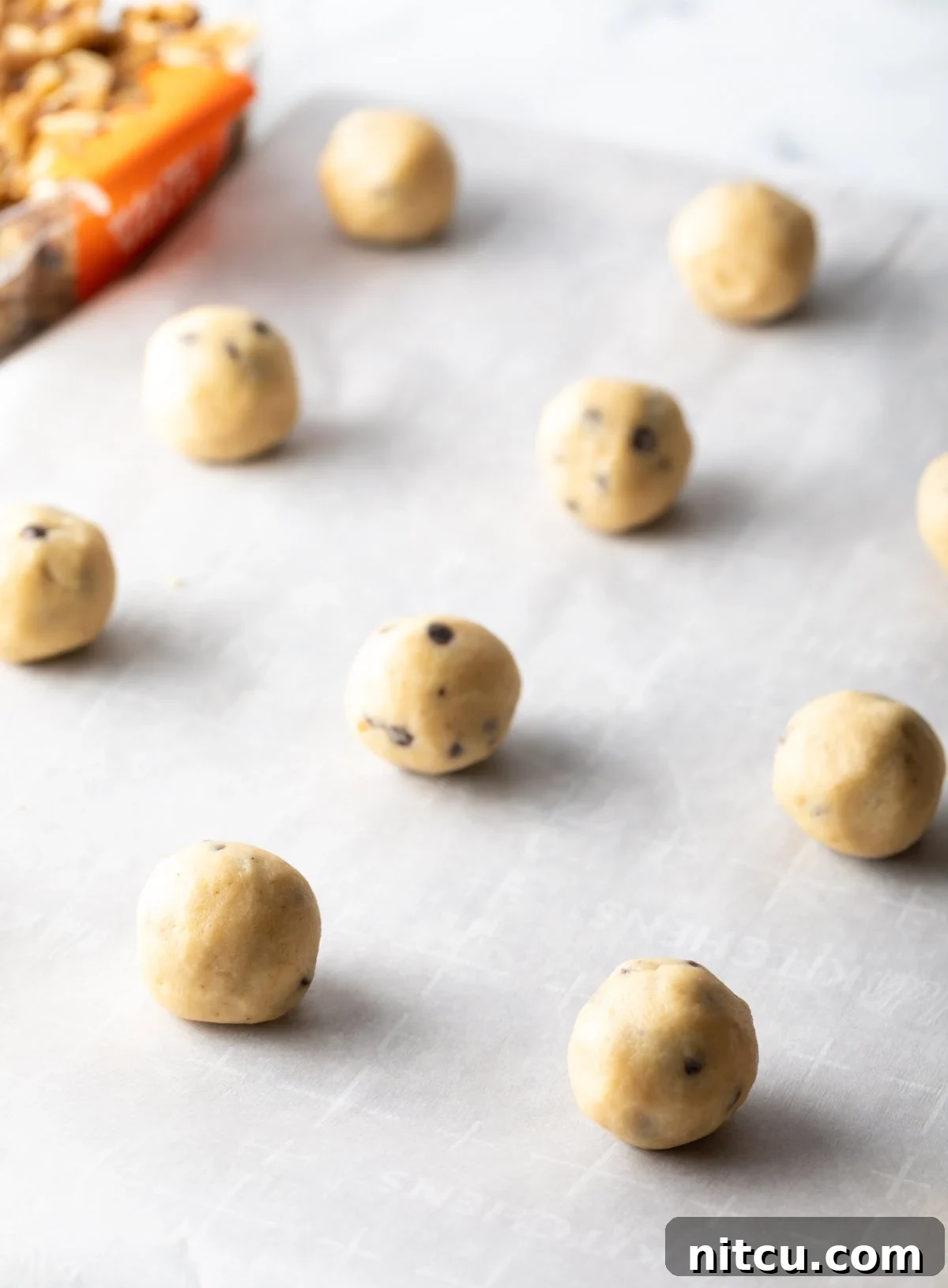 Chocolate chip walnut cookie dough formed into balls and lined up on a baking sheet with white parchment paper.