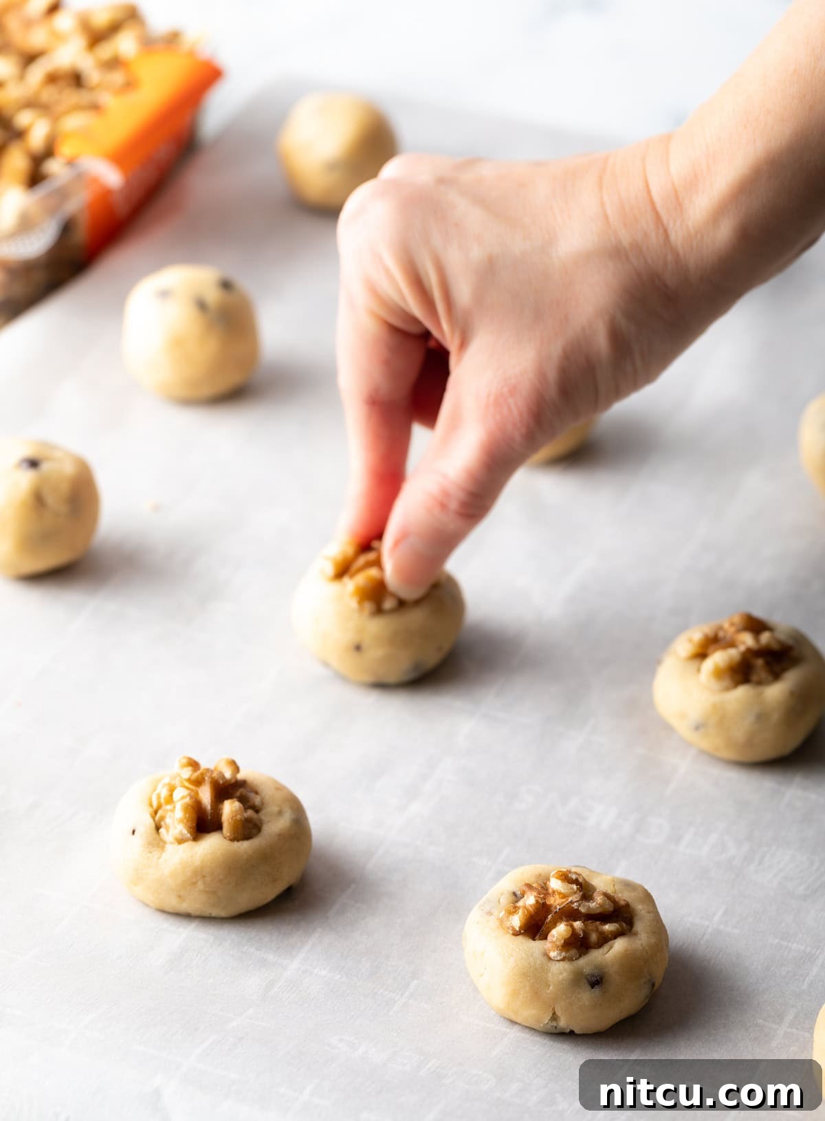 A hand is pressing a halved walnut into the balls of chocolate chip and walnut cookie dough.