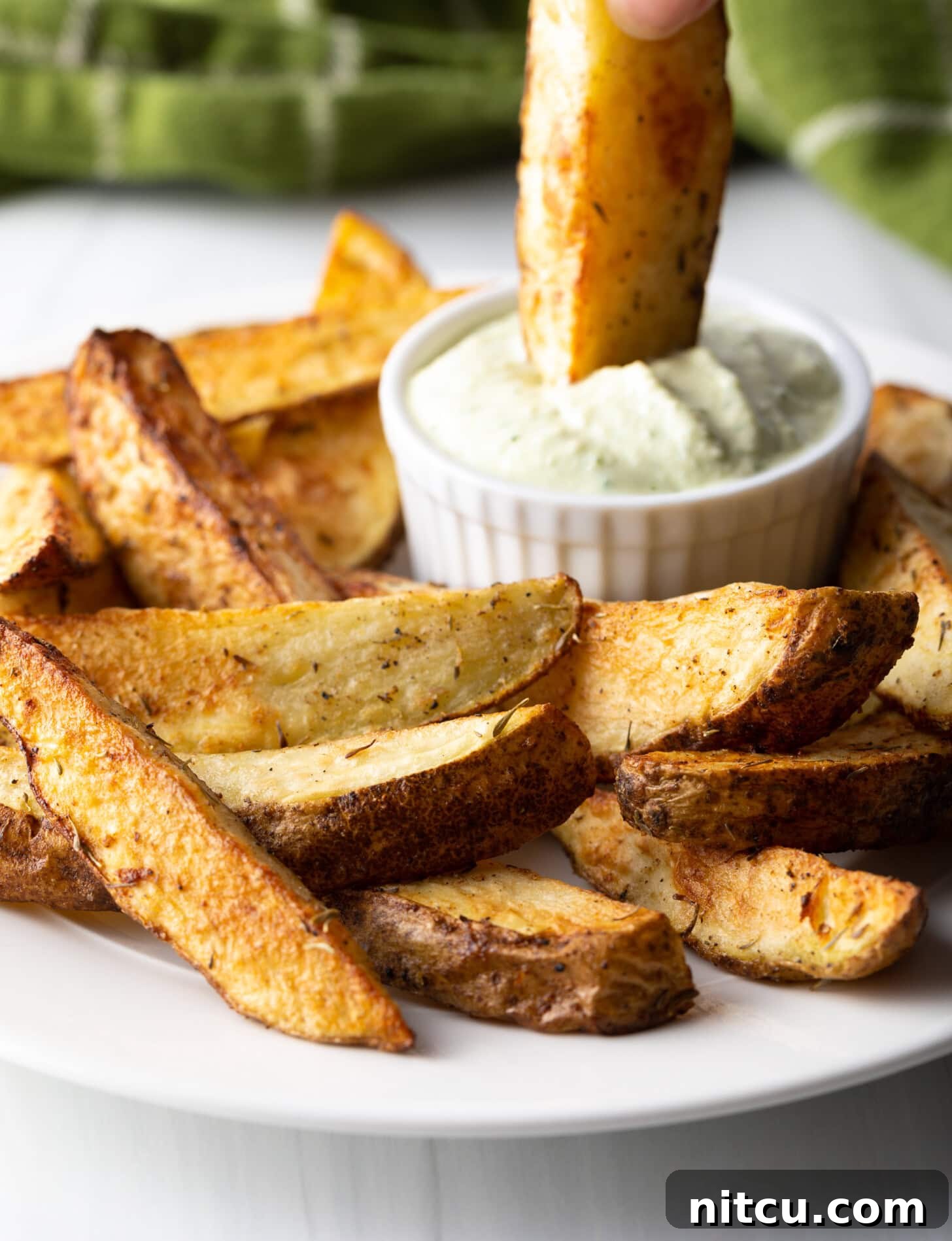 Crispy Golden Air Fryer Potato Wedges 11 A hand gently dips a golden-brown, crispy air fryer potato wedge into a small white ramekin filled with creamy white dipping sauce.