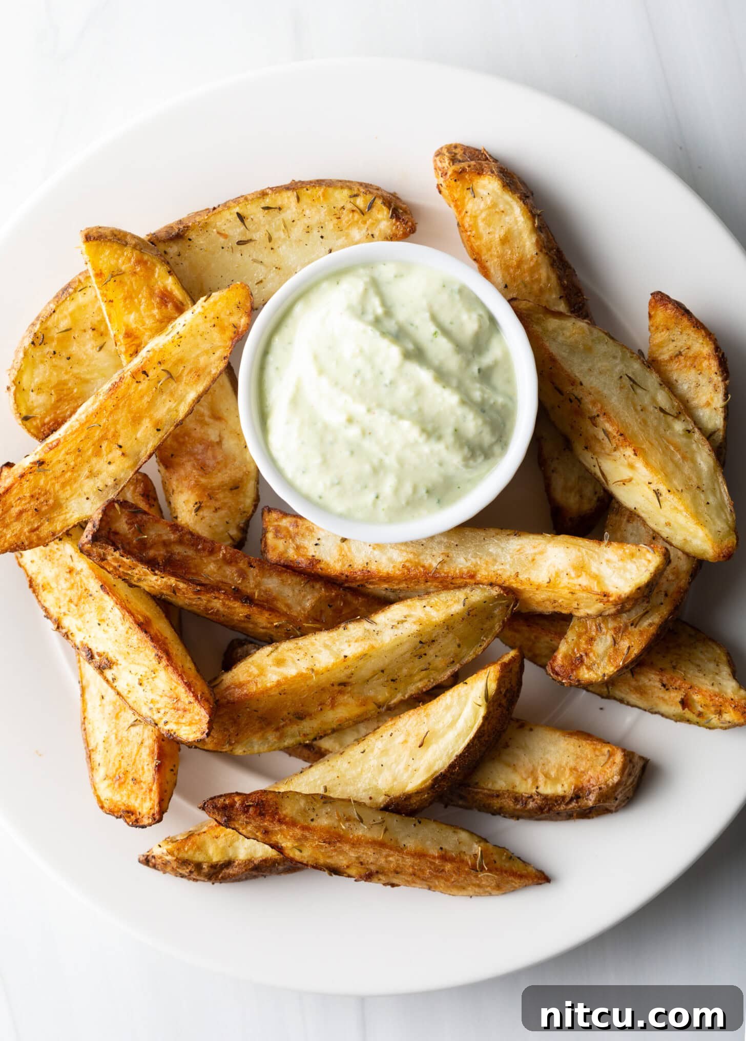 Crispy Golden Air Fryer Potato Wedges 12 A white plate is generously stacked with thick-cut, golden-brown air fryer potato wedges, accompanied by a small white ramekin holding a creamy dipping sauce.