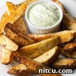 Crispy Golden Air Fryer Potato Wedges 14 A white plate piled with thick-cut air fryer potato wedges, with a white ramekin of white dipping sauce also on the plate.