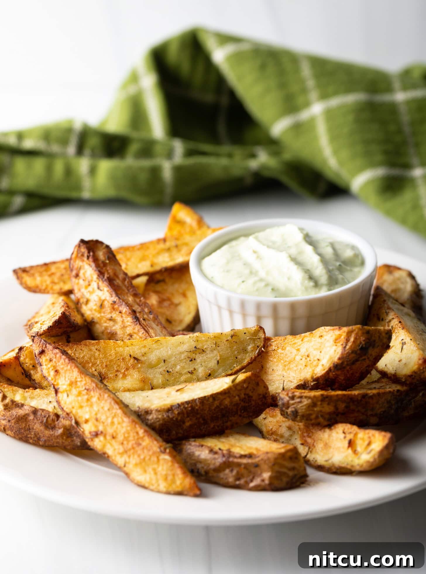 Crispy Golden Air Fryer Potato Wedges 3 A pristine white plate showcases a generous serving of crispy potato wedges, freshly made in the air fryer. Beside them, a small white ramekin holds a creamy, light-colored dipping sauce.