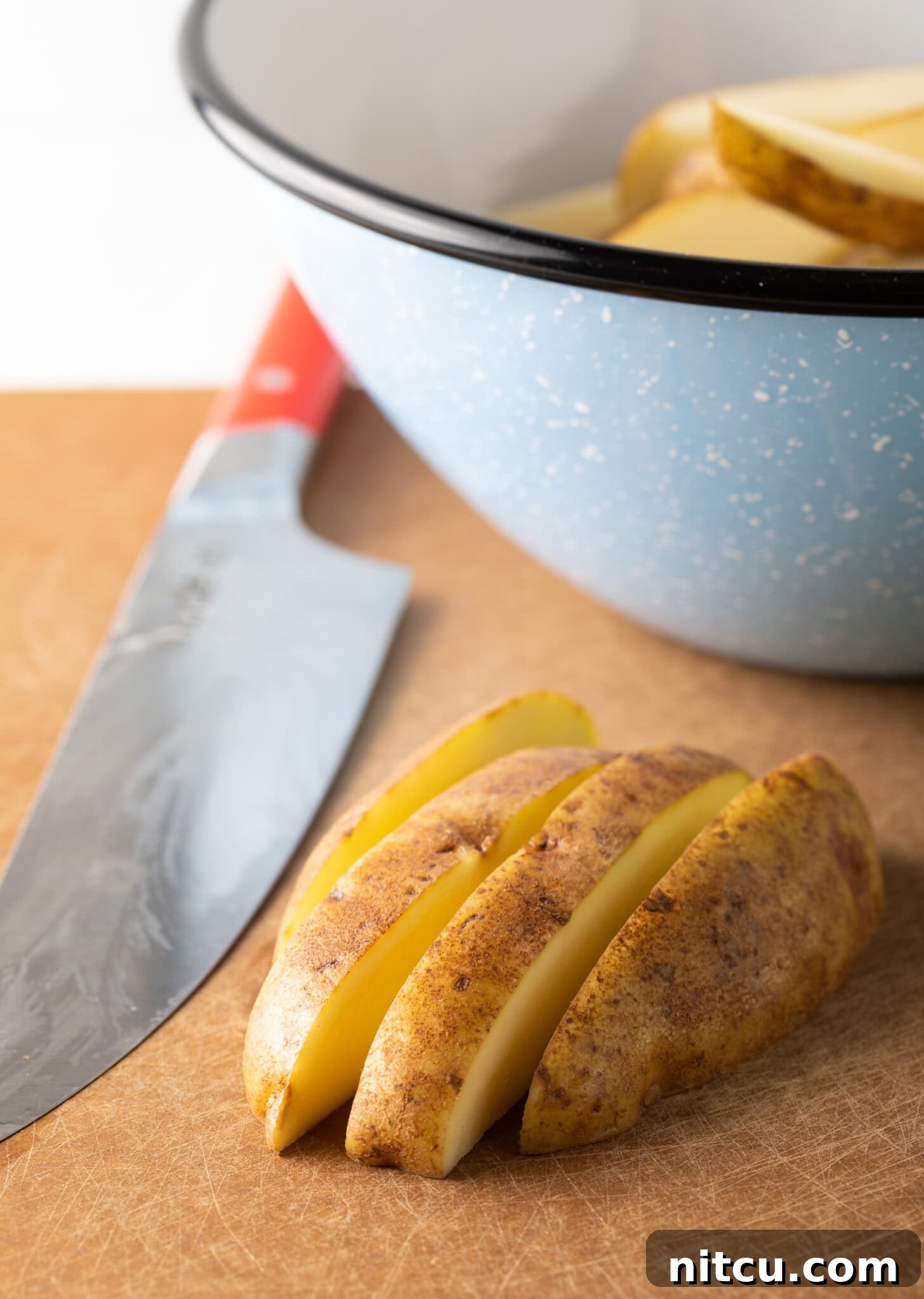 Crispy Golden Air Fryer Potato Wedges 6 A cutting board holds a half-cut raw potato, skillfully sliced into thick, uniform wedges with a sharp knife nearby.