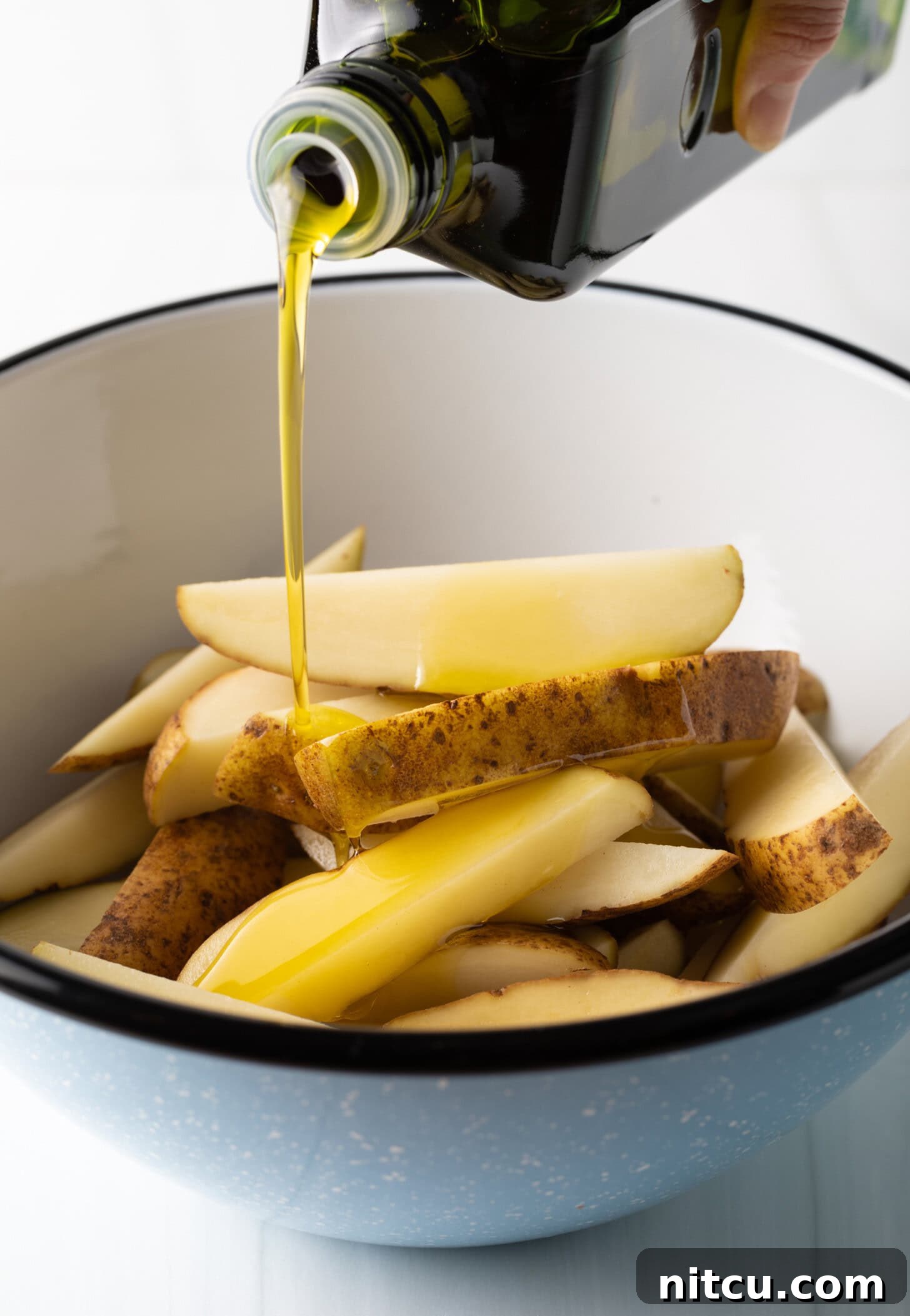 Crispy Golden Air Fryer Potato Wedges 7 A hand is captured in the act of pouring olive oil from a bottle directly onto thick-cut potato wedges in a pristine white bowl, preparing them for the air fryer.