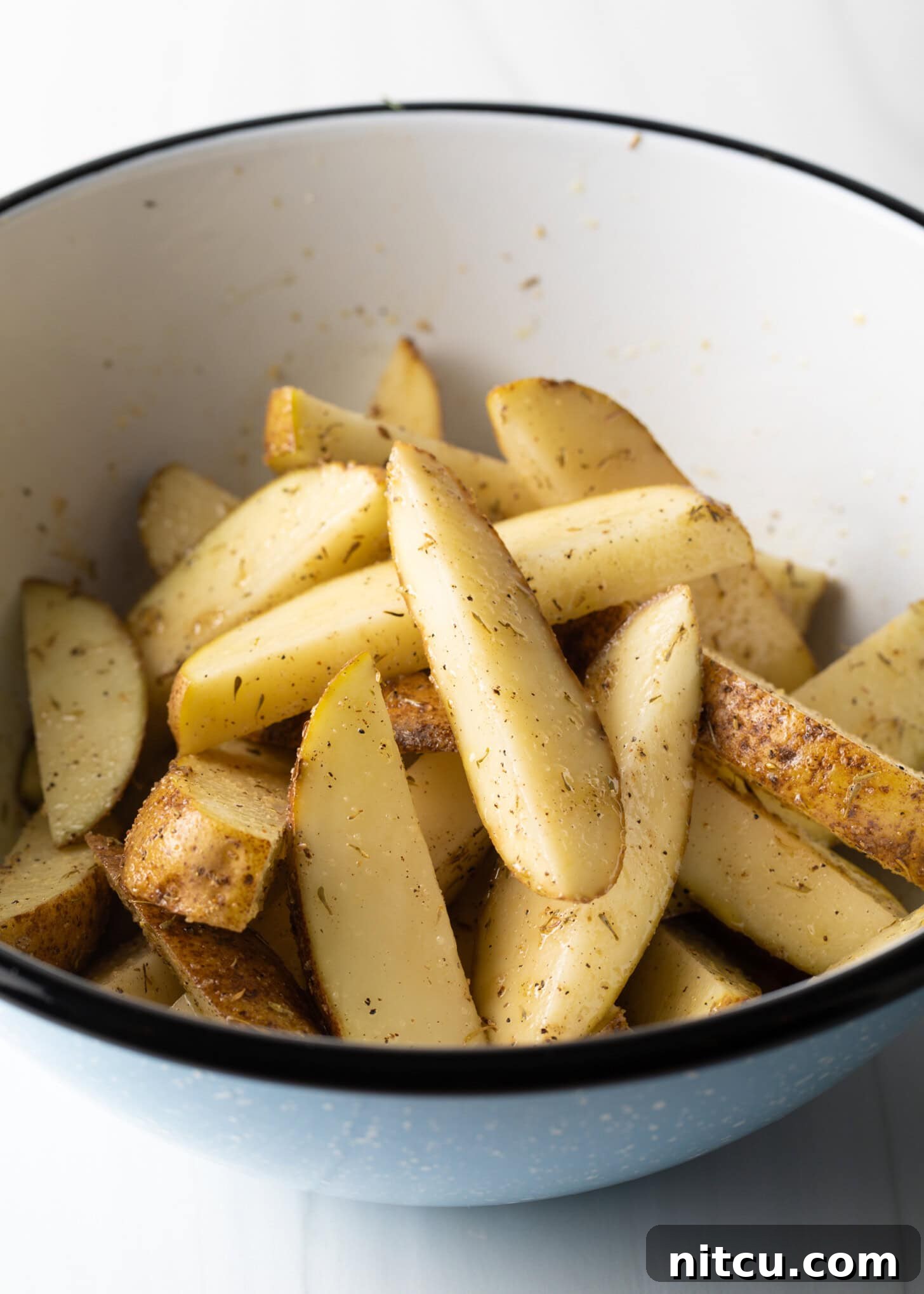 Crispy Golden Air Fryer Potato Wedges 8 Thick-sliced raw potato wedges are visible in a white mixing bowl, perfectly tossed with olive oil and a vibrant blend of seasonings.