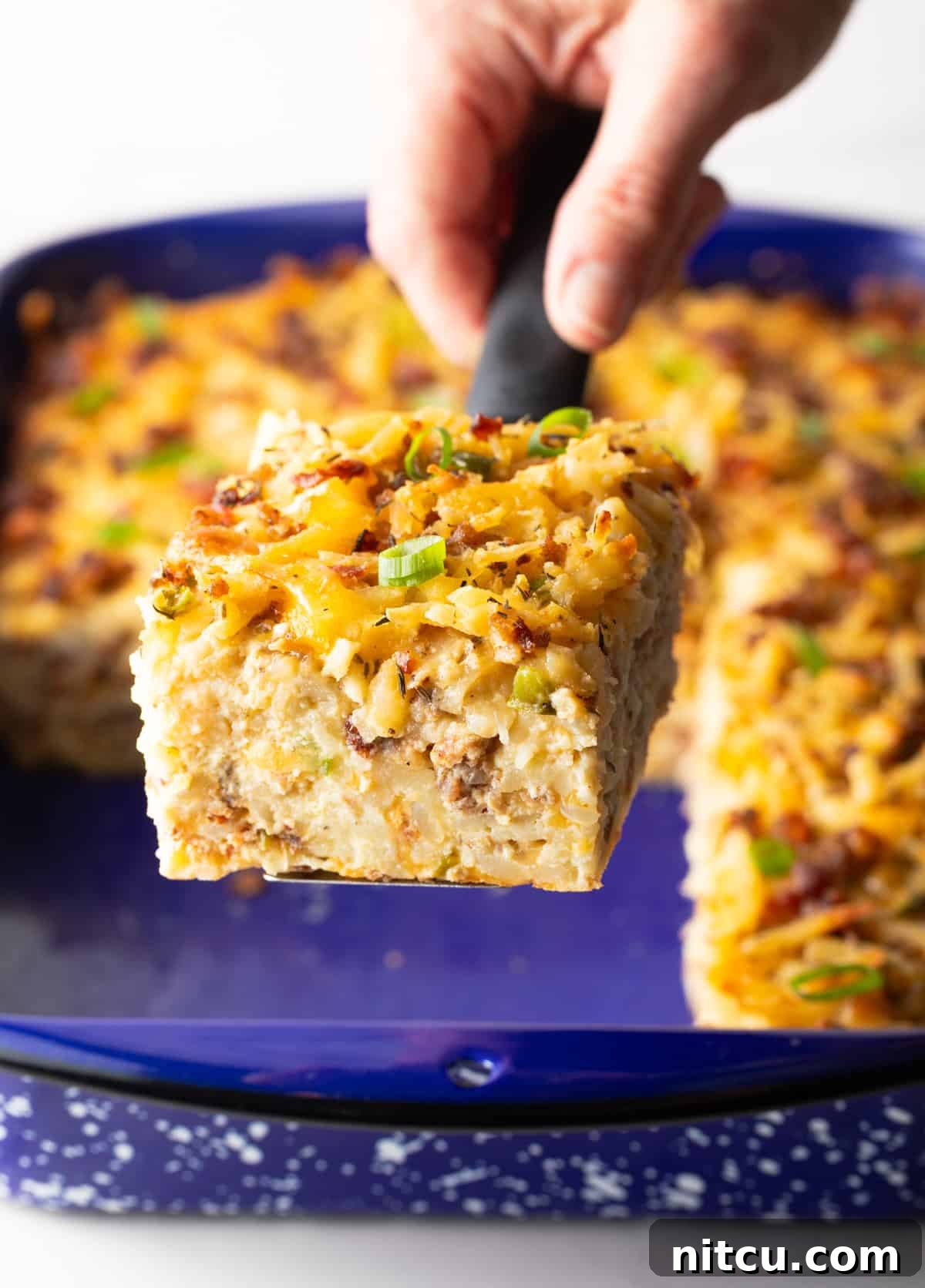 Golden Hash Brown Breakfast Bake 12 A hand holds up a perfectly square slice of golden-brown breakfast casserole towards the camera, highlighting its delicious texture.