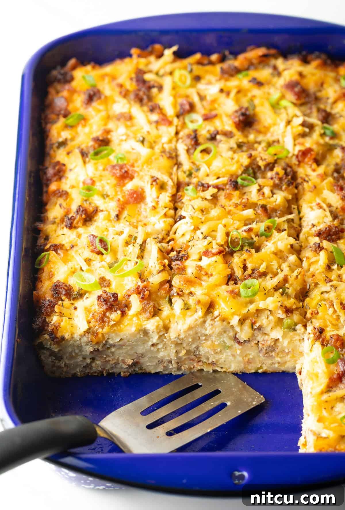 Golden Hash Brown Breakfast Bake 13 A freshly baked breakfast hash brown casserole, with two slices already served, is perfectly presented in a blue baking dish alongside a metal spatula.
