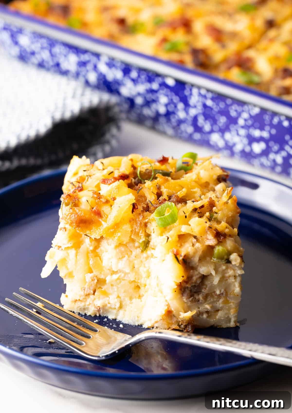 Golden Hash Brown Breakfast Bake 14 A delectable slice of breakfast casserole with hash browns rests on a blue plate, with a fork poised to take another tempting bite.