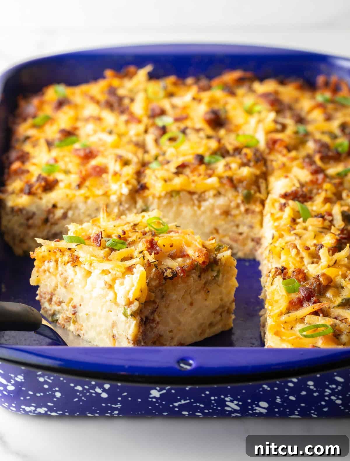 Golden Hash Brown Breakfast Bake 3 A full hash brown breakfast casserole in a vibrant blue baking dish, with a metal spatula lifting a single perfect slice.
