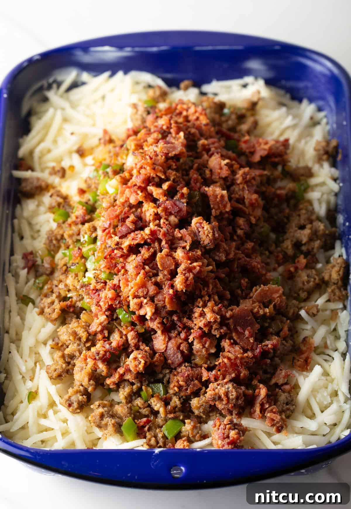 Golden Hash Brown Breakfast Bake 7 Frozen hash browns form the base layer, topped with a delicious mixture of cooked sausage and bacon, ready for the next step in a blue casserole dish.