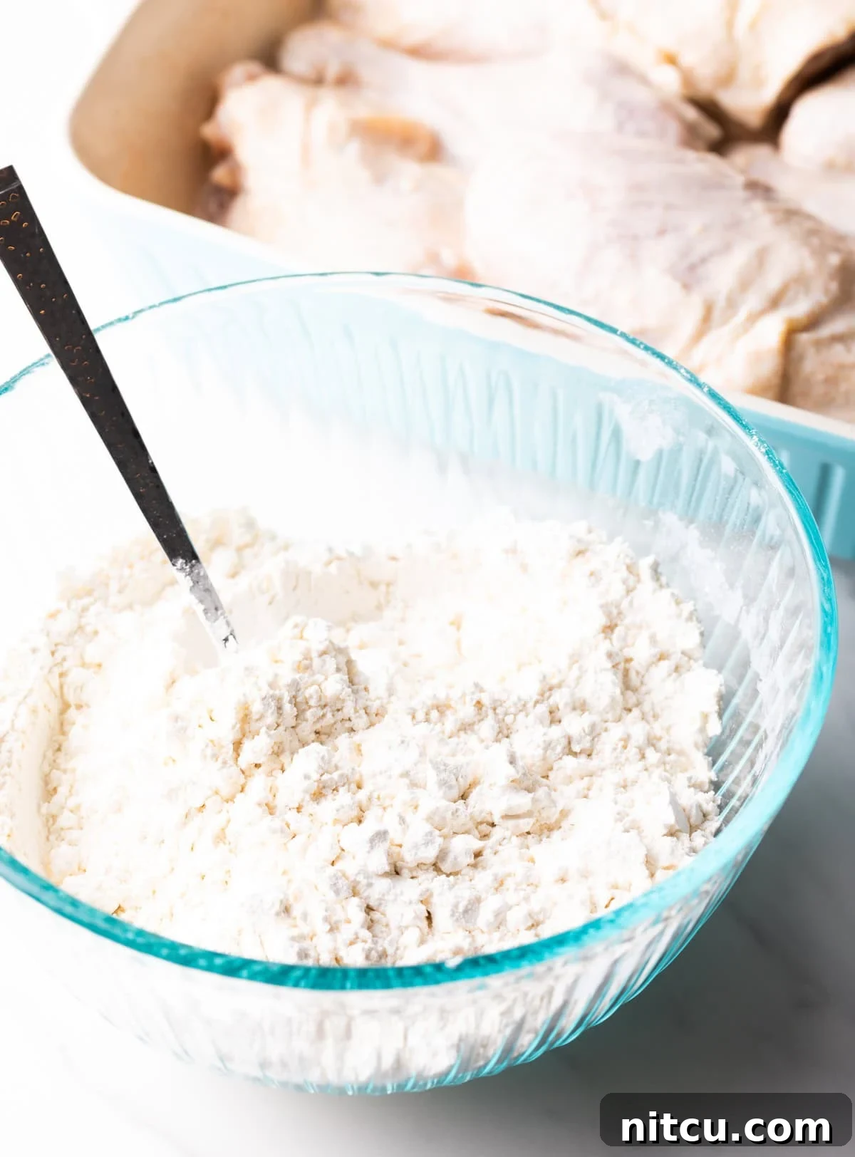 A spoon in a glass mixing bowl that is mixing a flour mixture for a deep fried chicken recipe.