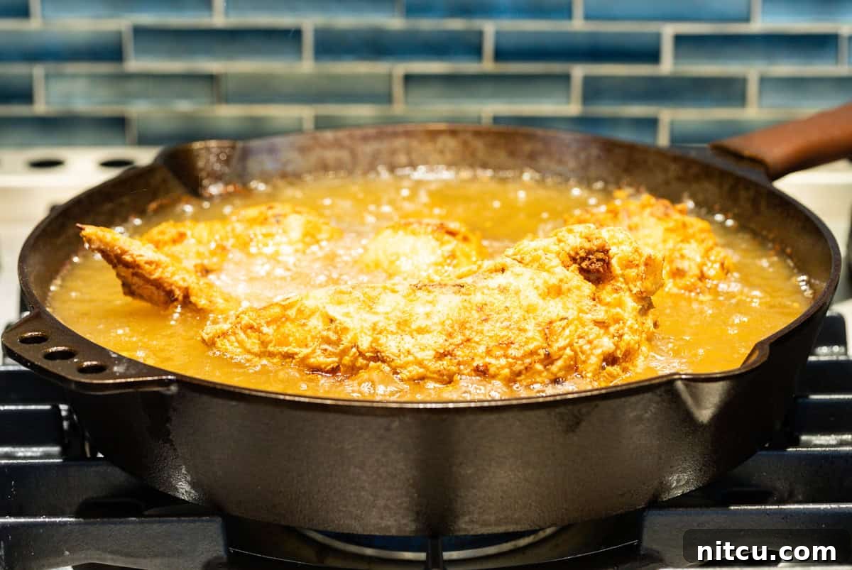 Fried chicken pieces are being cooked in a cast-iron skillet full of oil.