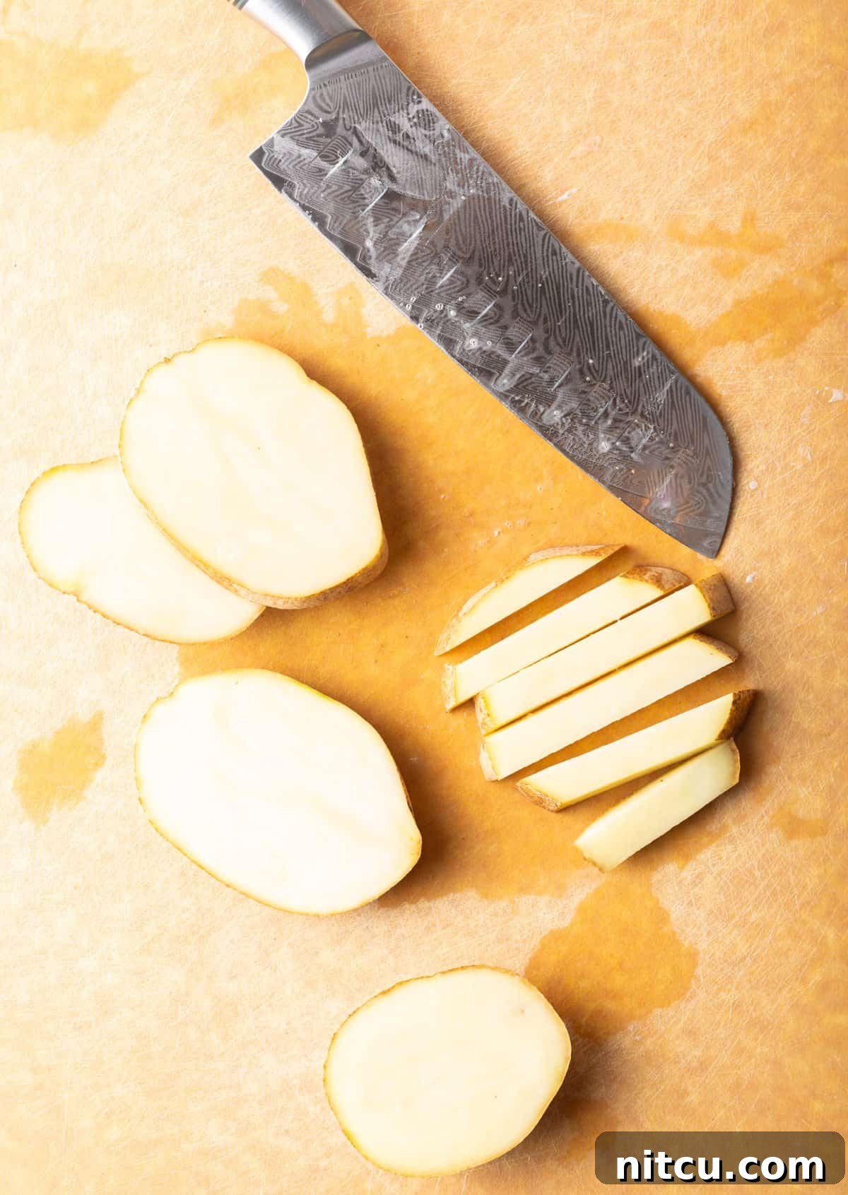Top down view slices and sticks of raw potato on a cutting board with a large knife.