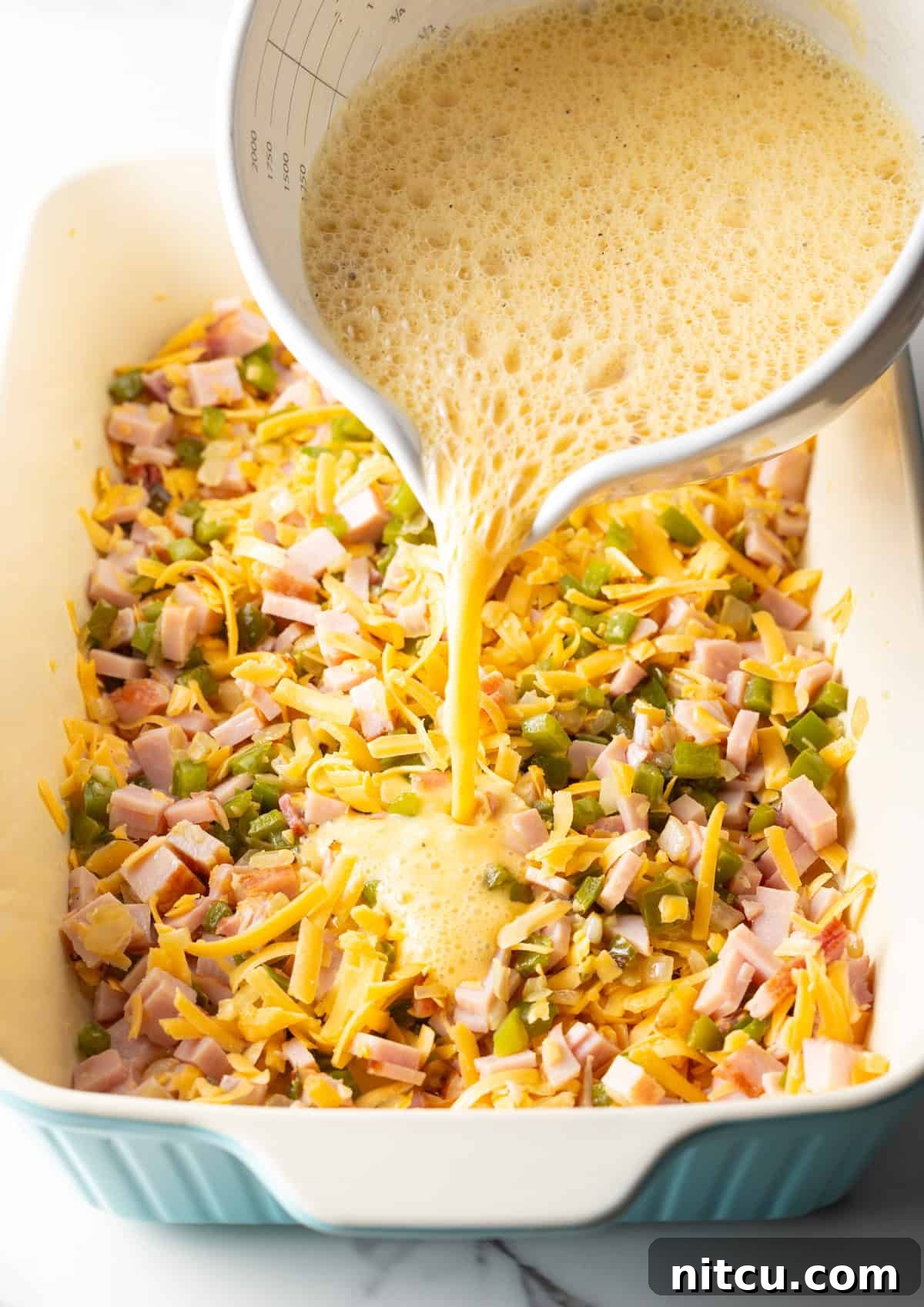 Mile-High Breakfast Casserole 11 An egg mixture is being poured over ham, cheese, and vegetables, in a white baking dish.