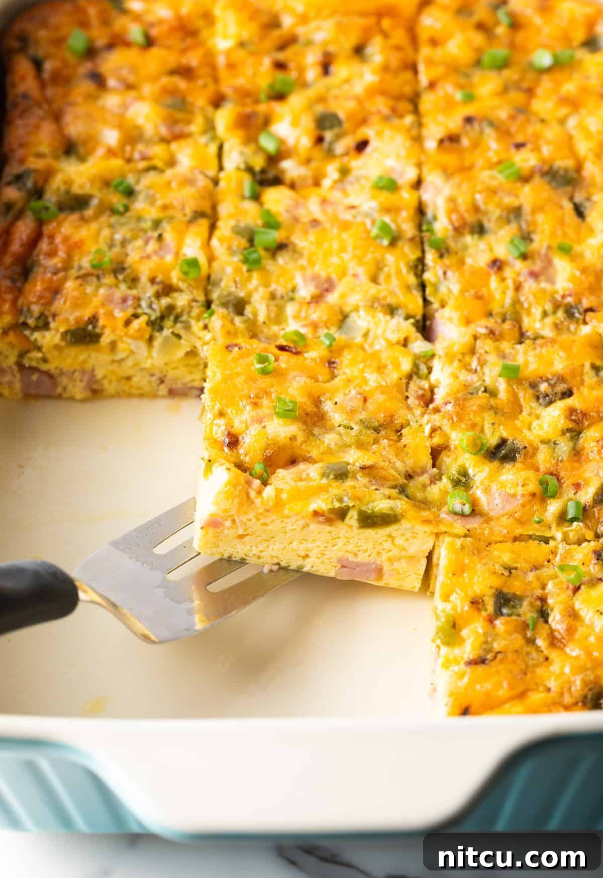 Mile-High Breakfast Casserole 13 A slice of a western omelette casserole is being scooped by a metal spatula over a white baking tray filled with a Denver omelette casserole.