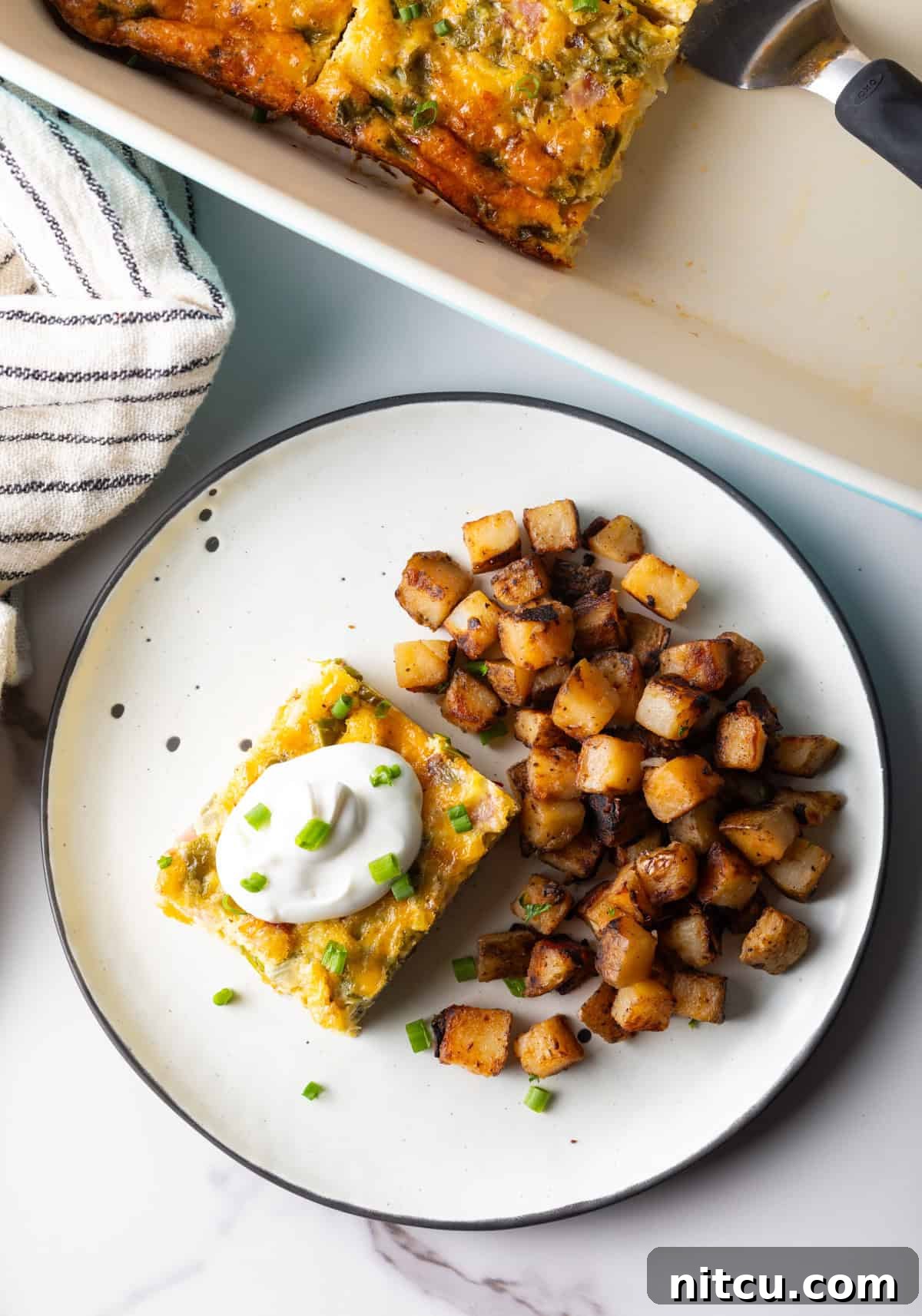 Mile-High Breakfast Casserole 14 An overhead shot of a slice of baked Denver omelet casserole topped with sour cream, next to a serving of home fries on a white plate.