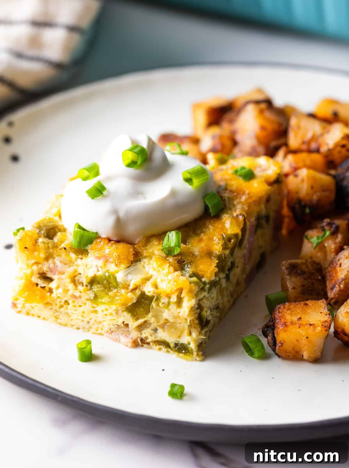Mile-High Breakfast Casserole 15 A slice of Denver baked omelet casserole topped with sour cream, next to a serving of home fries on a white plate.
