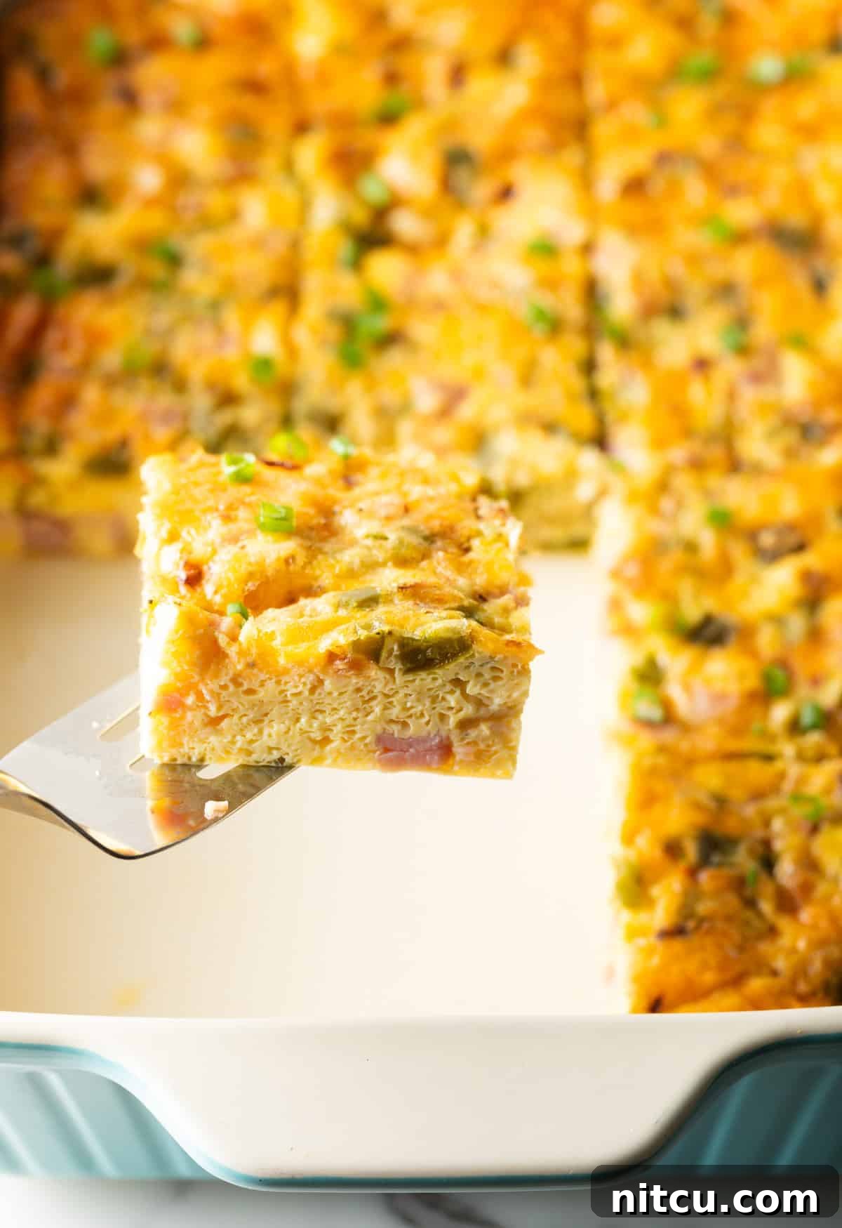 Mile-High Breakfast Casserole 5 A slice of a Denver baked omelette is being held by a metal spatula over a white baking tray filled with a Denver omelet casserole.