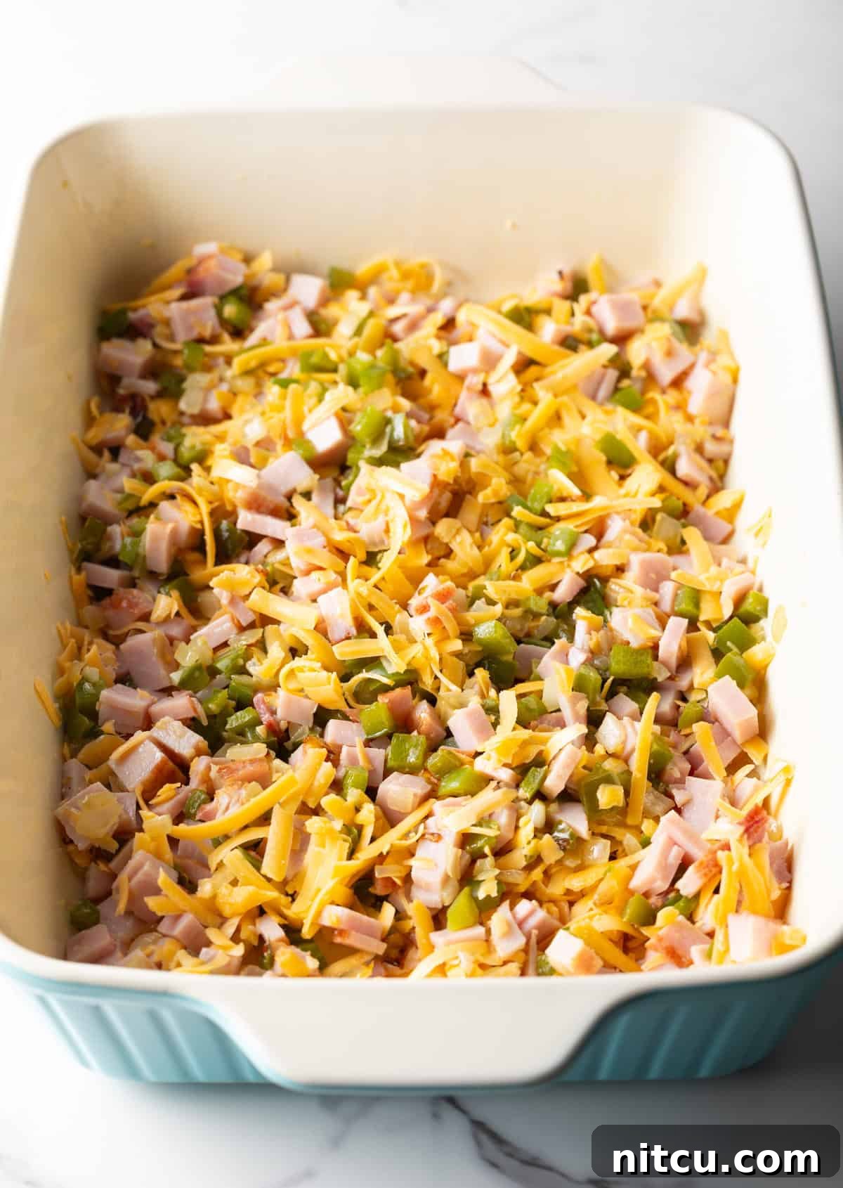 Mile-High Breakfast Casserole 10 Shredded cheese is being mixed into a ham and vegetable mixture in a white baking dish.